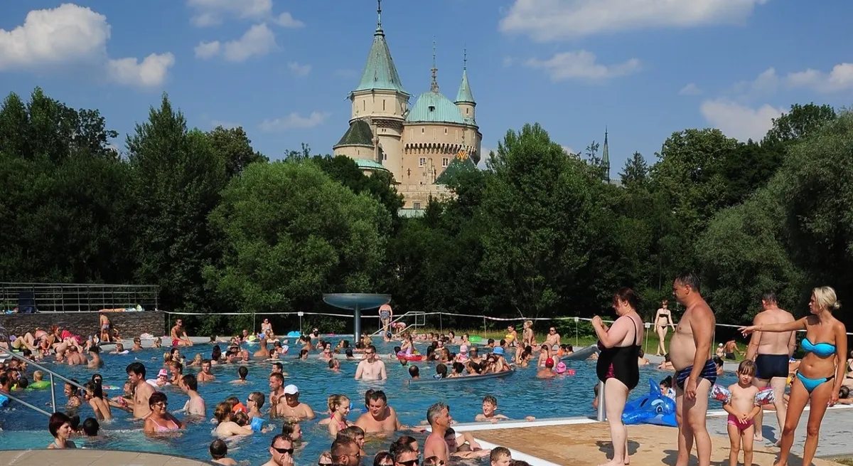 Ľudia, ktorí si užívajú teplý deň v termálnom kúpalisku Čajka v Bojniciach s výhľadom na Bojnický zámok.